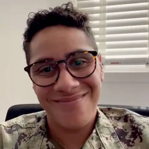 Rae Timberlake Rae sits in a chair wearing camouflage clothing and glasses. They have short dark hair and are smiling at the camera.