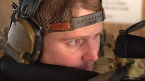 Close-up of Kara's face as she aims a rifle. She is wearing a back-to-front cap and ear defenders.