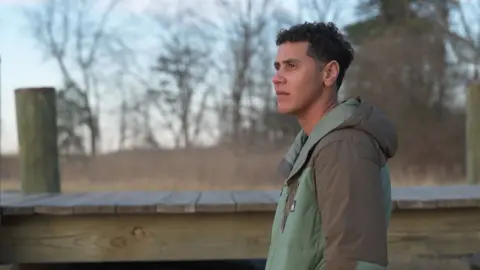 Rae looks pensive with a jetty and trees in the background. They are wearing a green and beige hooded jacket. 