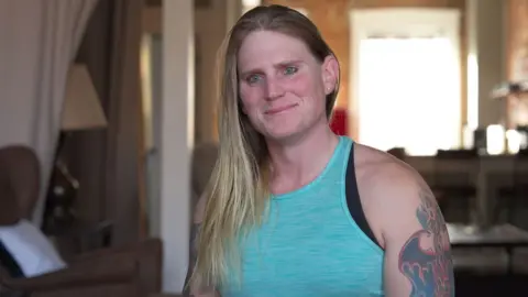 BBC Kara's long straight blonde hair falls over her right shoulder. She is wearing a light blue vest top and has a tattoo of a phoenix rising, engulfed in flames, on her left arm.